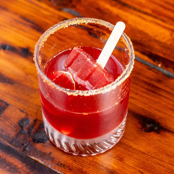 Red cocktail with a sugar-rimmed glass, garnished with a red popsicle and lime slice.