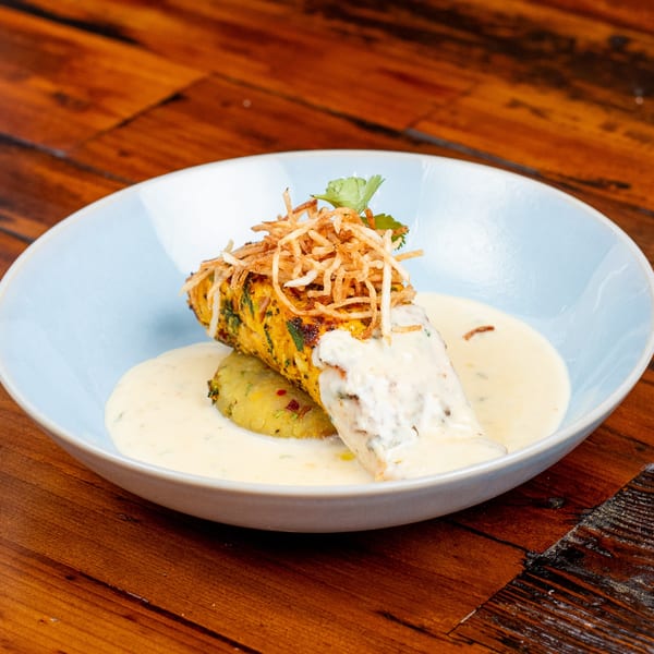 A light blue bowl holds a golden-brown fish fillet with crispy toppings, a potato cake, and creamy sauce.