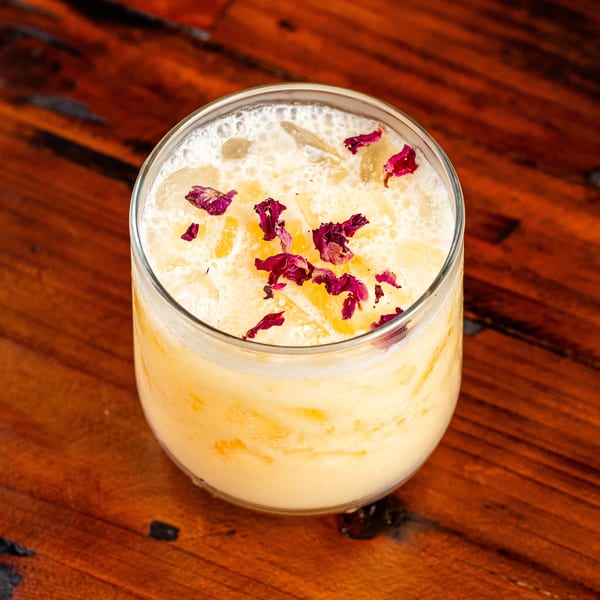 Creamy yellow cocktail with ice and dried rose petals, on a dark wooden table.