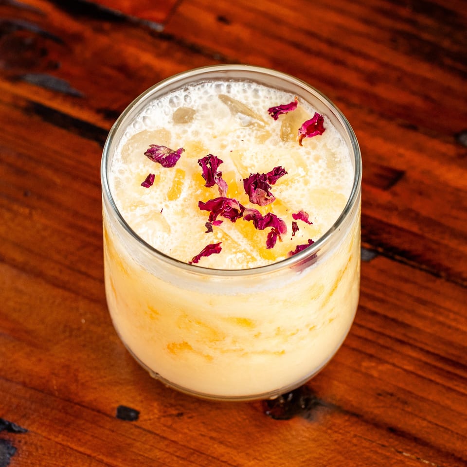 Creamy yellow cocktail with ice and dried rose petals, on a dark wooden table.