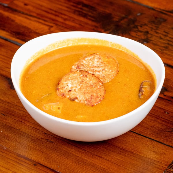 A white bowl of orange soup with two round, crispy hash browns on top, on a wooden table.