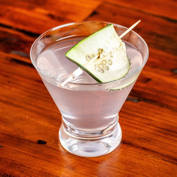 Light pink martini in a glass, garnished with a cucumber slice on a wooden table.