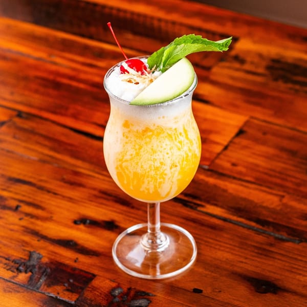A vibrant yellow cocktail with white foam, garnished with a cherry, mint, and melon slice on a wooden table.