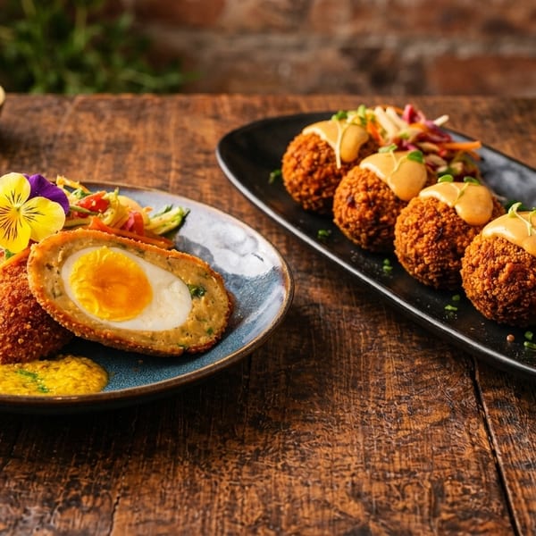 Two plates of Scotch eggs, one halved with a runny yolk, on a rustic wooden table.