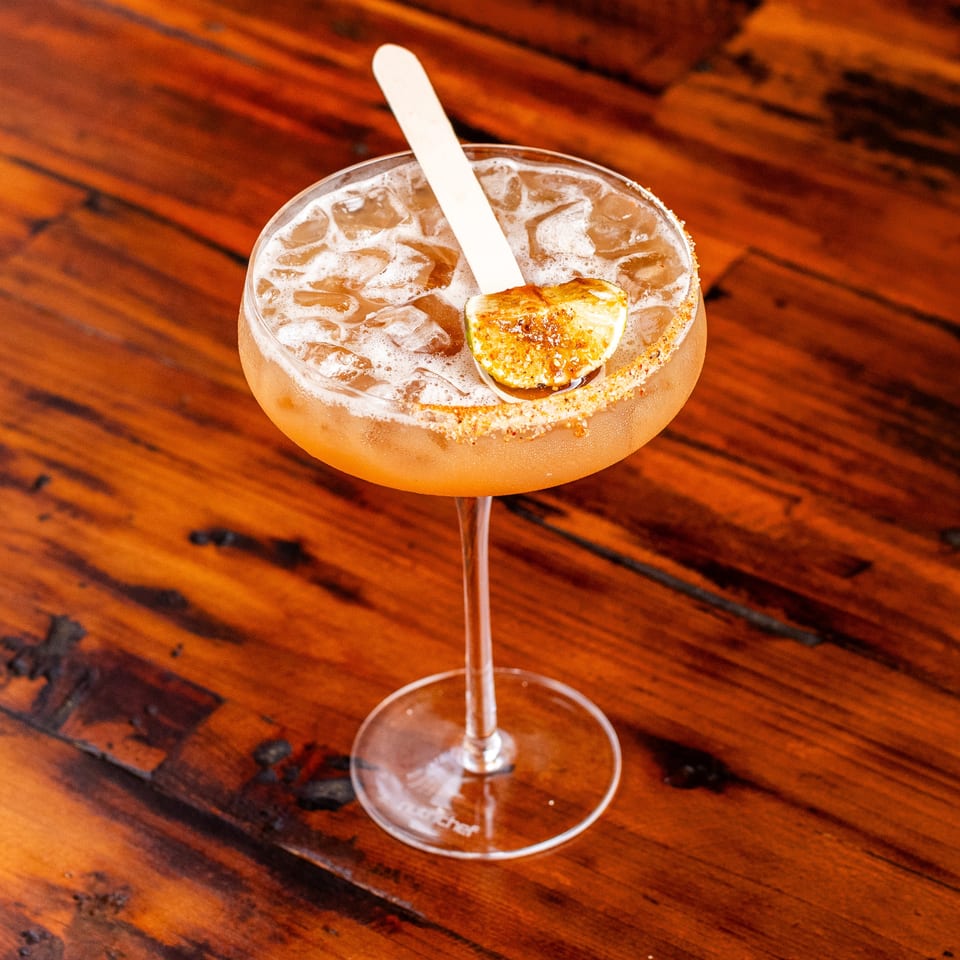 Iced cocktail with a sugared rim, garnished with a lime wedge and a wooden stirrer on a wooden table.