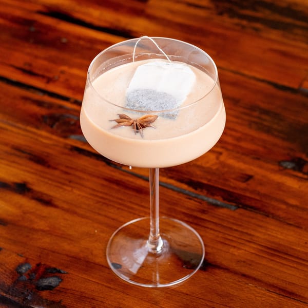 Chai cocktail with a tea bag and star anise garnish in a stemmed glass on a wooden table.