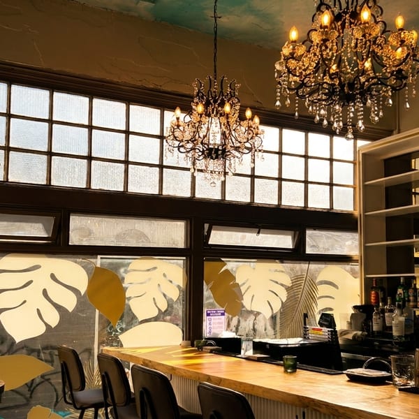 A bar interior with two ornate chandeliers, a long wooden bar, and large windows with leaf decals.