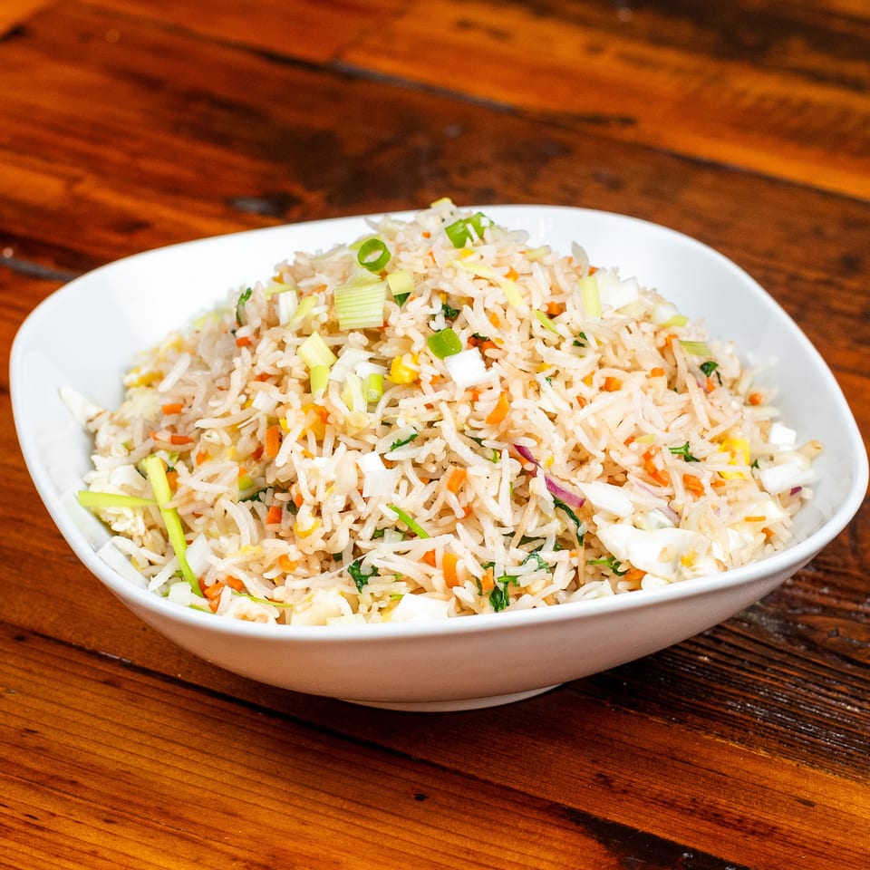 A white bowl filled with vegetable fried rice, garnished with chopped green onions, on a wooden table.