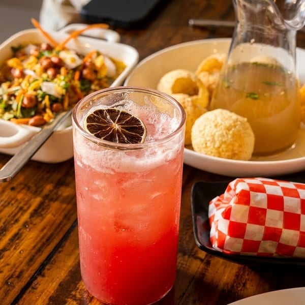 A vibrant red cocktail with a dried lime slice, surrounded by various appetizers on a wooden table.